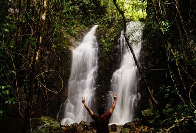 Rear view of man with arms raised against waterfall in forest