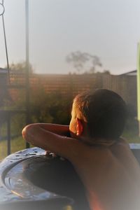 Rear view of shirtless boy leaning on wet railing in back yard against sky during sunset