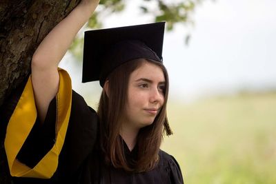 Thoughtful young woman wearing graduation gown and mortarboard by tree trunk