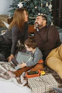 Family with child playing near christmas tree. child unpacking gifts, parents enjoy christmas tree