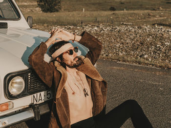 Serious male hippie in outerwear and headband sitting near white old timer automobile during trip in countryside on sunny day