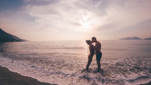 Side view of couple kissing while standing on shore at beach against sky