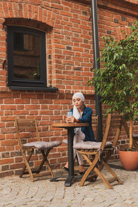 Portrait of smiling young woman sitting on chair against brick wall