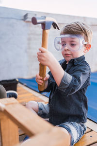 Child in denim shirt and plastic glasses sitting with hammer against wooden piece in daylight