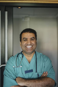 Portrait of cheerful mature doctor standing with arms crossed in front of door at hospital