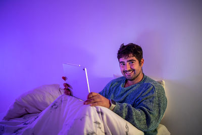 Portrait of man using digital tablet sitting on bed against wall at home