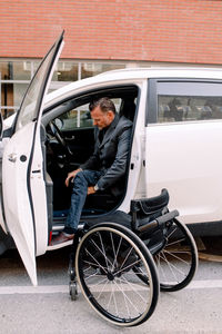 Full length side view of mature disable man disembarking from car with wheelchair on street in city