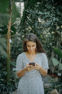 Young woman holding smart phone while standing by plants