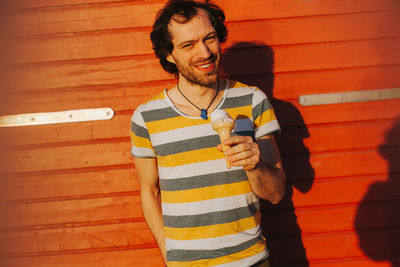 Portrait of smiling man holding ice cream cone while standing against wall