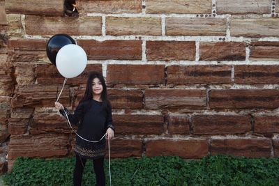 Portrait of smiling girl holding balloons while standing against brick wall