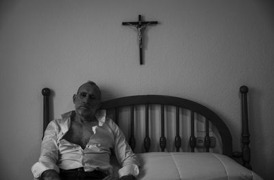 Monochrome of adult man in white shirt in room lying on bed with religion cross on wall
