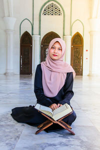 Full length portrait of young woman reading holy book while sitting at mosque