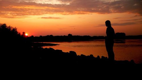 Silhouette man standing by lake against orange sky