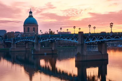 Bridge over river by buildings against sky during sunset