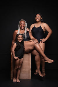 Full length of multiethnic plus size females of various ages wearing black bodysuits smiling and looking at camera in studio with wooden bench and black background