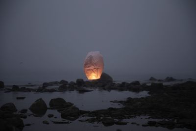 Paper lantern flying over rocks at beach against sky during dusk