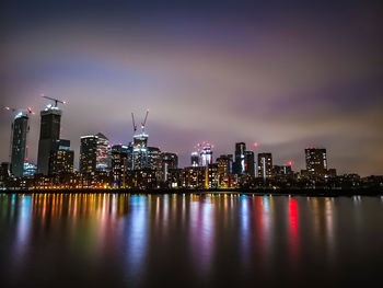 Illuminated buildings by river against sky at night