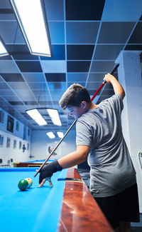 Side view of serious boy in casual wear with cue stick preparing to hit ball while playing pool game at blue billiard table