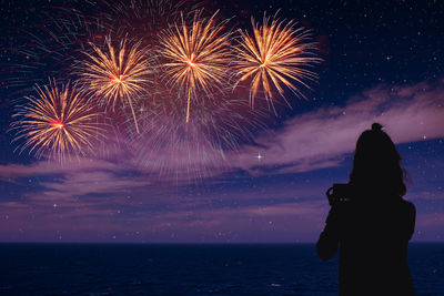 Rear view of woman standing against firework display over sea against sky at night
