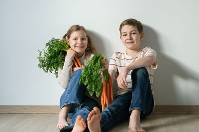 Portrait of smiling sibling holding carrot while sitting against wall