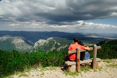 Rear view of couple sitting on wooden bench while looking at mountains against cloudy sky