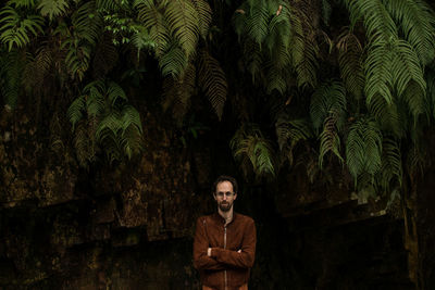 Portrait of young man standing with arms crossed on plant against trees