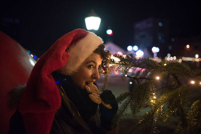 Young woman eating cookie by illuminated christmas tree at night