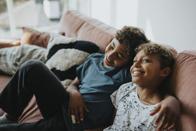 Happy boy with arm around brother sitting on sofa at home