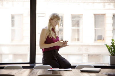Businesswoman using smart phone while sitting on window sill in board room