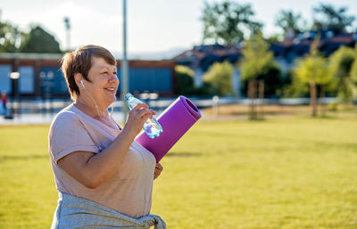 Happy smiling senior plus size woman with earphones holding yoga mat outdoors drinking water 