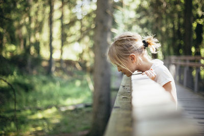 Side view of girl looking down while standing on footbridge in forest