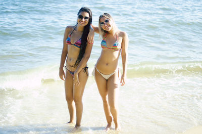 Full length portrait of smiling friends wearing bikini and sunglasses standing on shore at beach