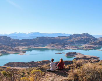 Rear view of woman sitting on rock against clear blue sky
