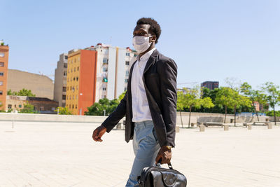 Well dressed black businessman in protective medical mask walking along street on sunny day in city during covid 19 epidemic and looking away