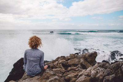 Rear view of woman sitting on rock by sea against sky