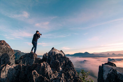 Man photographing while standing on rock against sky