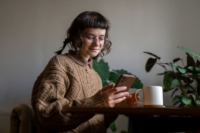 Pleased hipster woman drinking tea looking at smartphone screen smiling sitting at table at home.