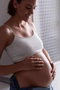 Side view of charming pregnant female smiling and gently touching tummy while standing in room at home