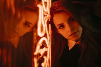 Young content female leaning on glowing orange neon sign and looking at camera while standing on street at night