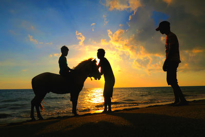 Silhouette people with horse at beach against sky during sunset