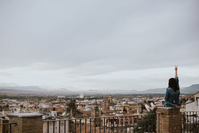 Rear view of woman gesturing peace sign while looking at cityscape against sky