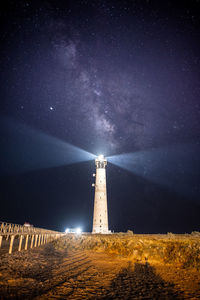 Lighthouse amidst illuminated buildings against sky at night