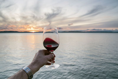 Point of view hand swirling red wine under a swirling cloud sunset over sea.
