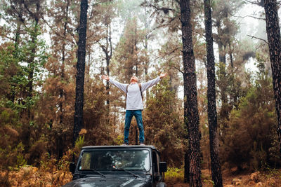 Full length of boy with arms outstretched standing on car at forest