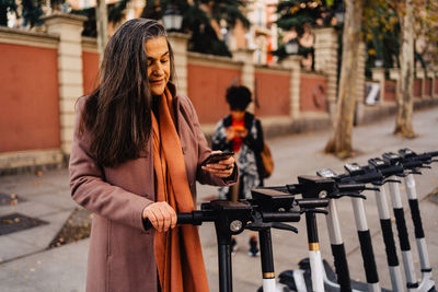 Hispanic long haired lady in warm coat standing on city street and doing contactless payment for electric scooter with smartphone using application