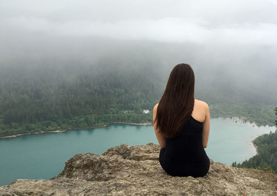 Rear view of woman sitting by lake against sky during foggy weather
