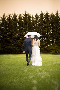 Rear view of newlywed couple walking on field while holding umbrella
