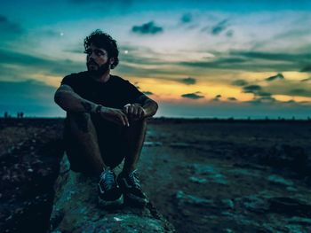 Full length of young man sitting on land against sunset sky