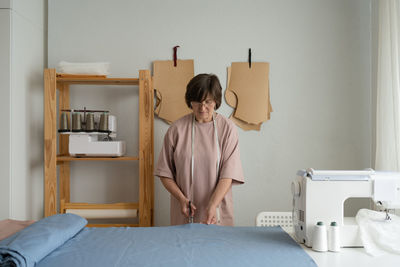 Concentrated professional female dressmaker with measuring tape cutting blue textile with scissors on table while working in modern light workshop