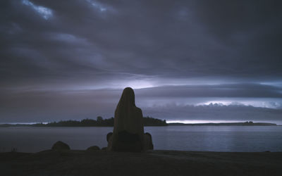 A woman in a headscarf sits on the shore and sees an approaching thunderstorm. night time. back view